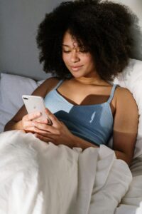 African American woman enjoying a cozy morning in bed, using a smartphone.